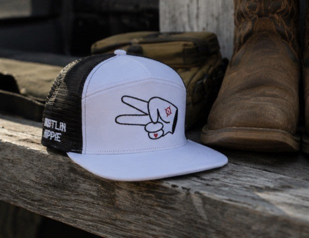 White and black Hustlin Hippie mesh trucker snapback hat with peace sign logo sitting on a rustic wooden bench beside cowboy boots and outdoor gear.
