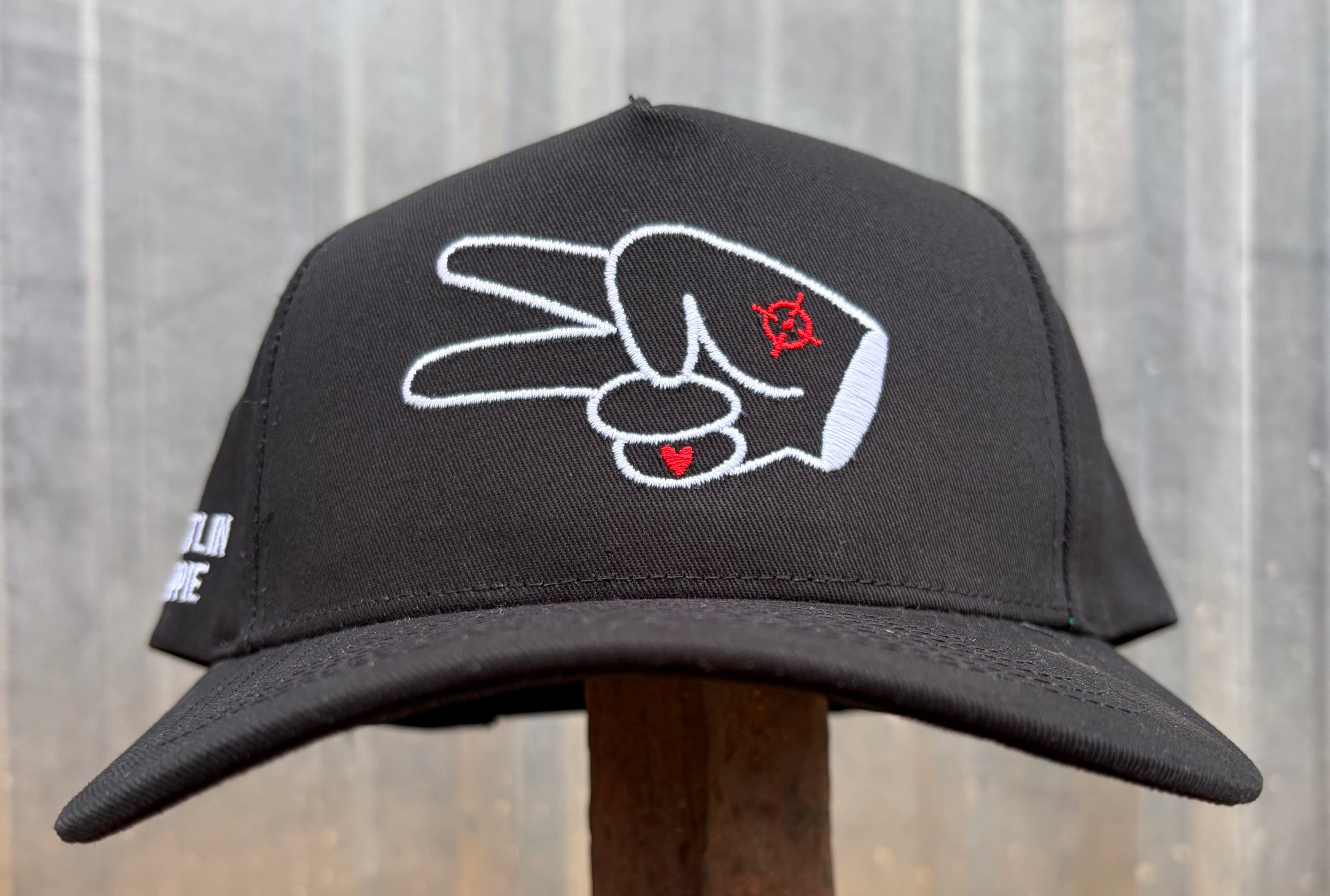 Black Hustlin Hippie curved bill snapback hat with embroidered signature peace hand logo on the front panel displayed outdoors on rustic metal stand