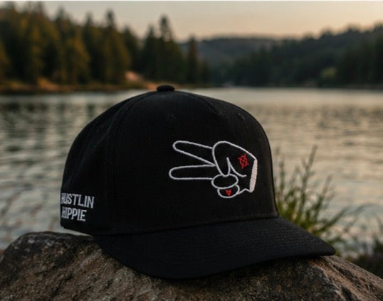 Black Hustlin Hippie curved snapback hat with embroidered peace hand logo displayed on a rock beside a lake at sunset with forest and water background outdoors