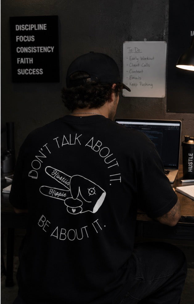 Man wearing black Hustlin Hippie motivational graphic t-shirt while working late at a desk in a dark modern workspace