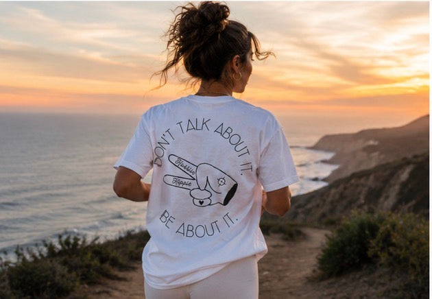 Woman wearing white Hustlin Hippie graphic t-shirt overlooking an ocean sunset on a coastal trail