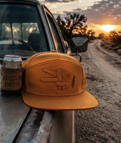 Golden orange Hustlin Hippie hydro snapback resting on a truck beside a dusty backroad at sunset with hunting and camping aesthetic.