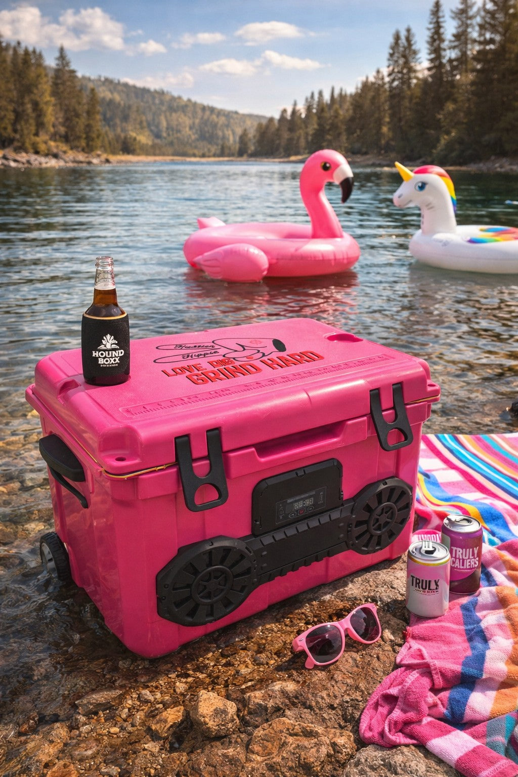 Pink Hound Boxx cooler with Bluetooth speaker on a lakeside with inflatable pool toys and summer accessories.