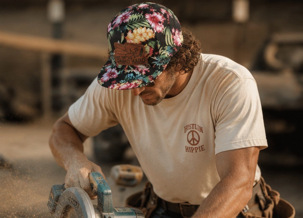 Man wearing floral Love Big Grind Hard leather patch snapback hat using circular saw on construction jobsite wearing Hustlin Hippie shirt