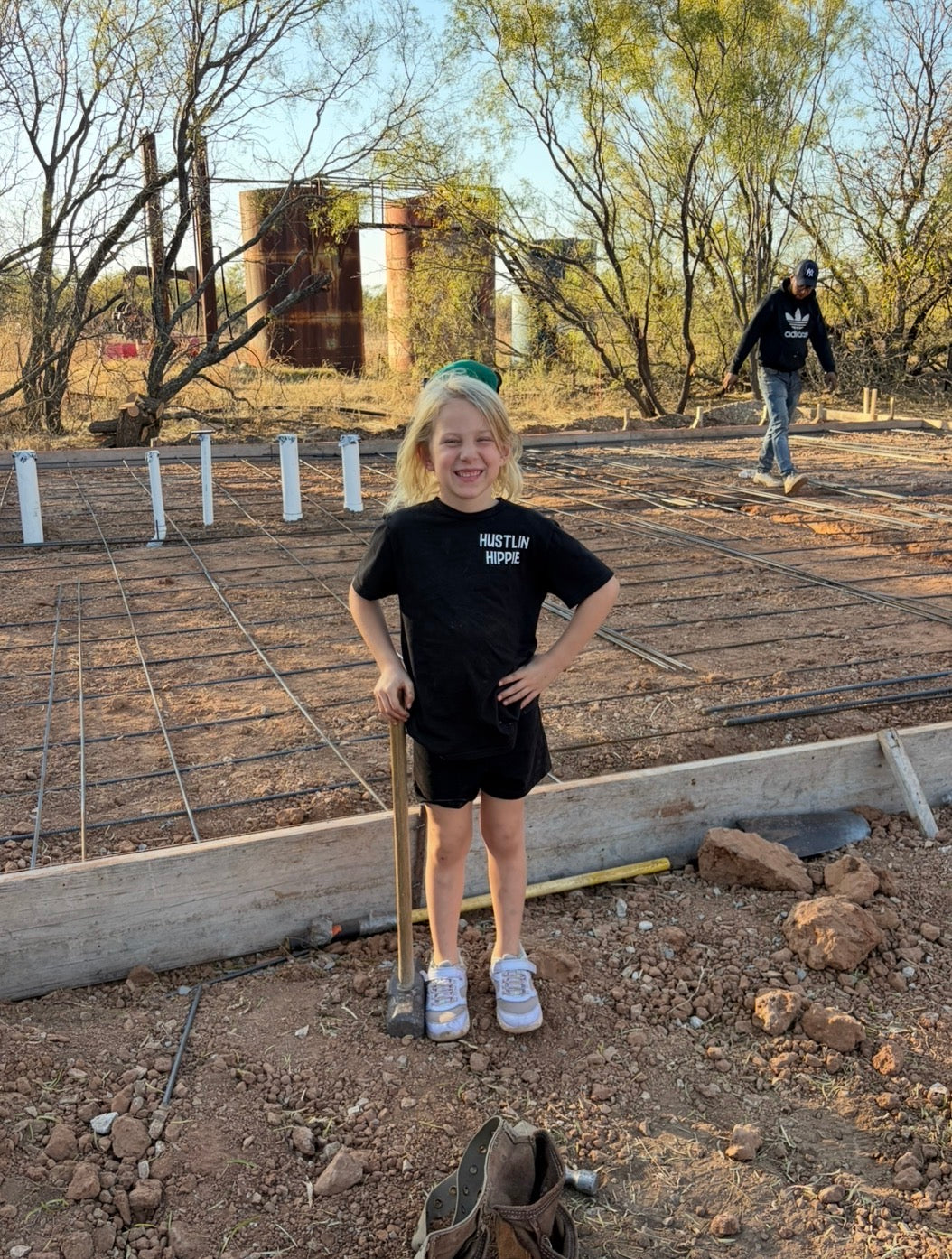 Child modeling black Hustlin Hippie Love Big Grind Hard youth t-shirt outdoors at a construction site.
