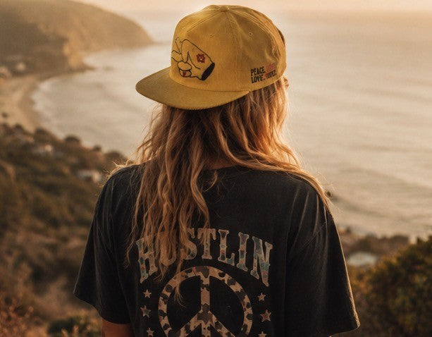 Person wearing a mustard yellow Hustlin Hippie corduroy snapback hat watching a canyon sunset with retro Good Vibes graphic tee