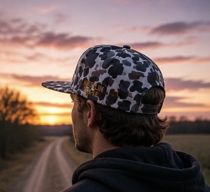 Rear lifestyle shot of a person wearing the Hustlin Hippie Signature Midnight Camo snapback on a country road at sunset.