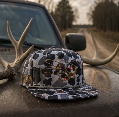 Hustlin Hippie Signature Midnight Camo rope snapback hat resting on a truck hood beside antlers during golden hour.