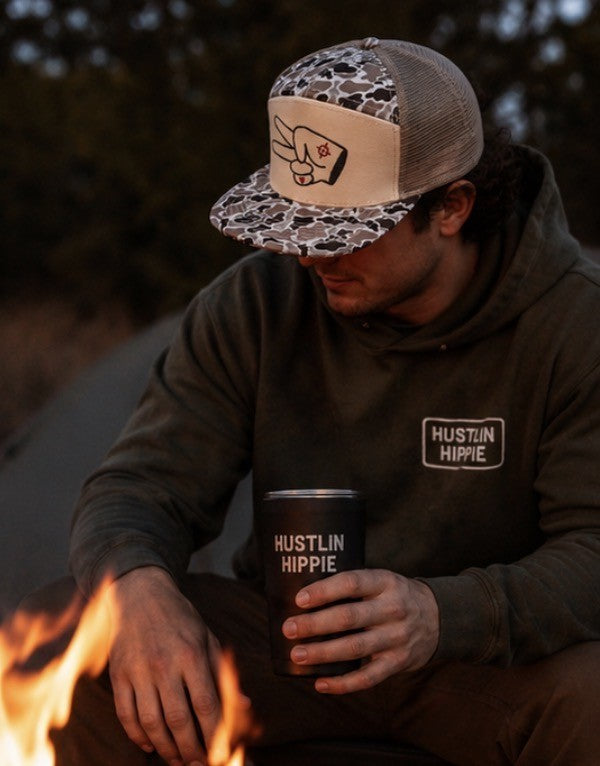 Man wearing Hustlin Hippie Signature Tan Camo Mesh Back Trucker Hat beside a campfire outdoors