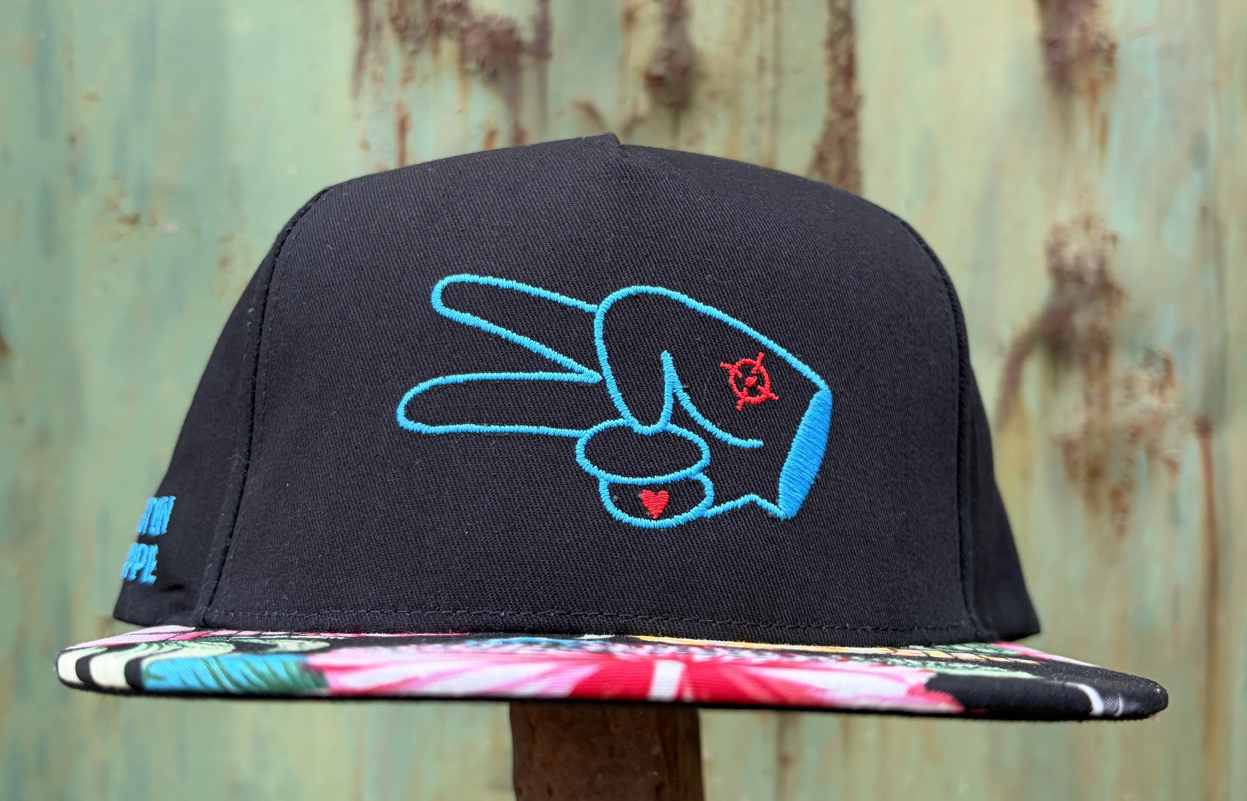 Close-up of the Hustlin Hippie Signature Tropical Floral flat bill hat featuring colorful floral artwork and embroidered peace hand logo.