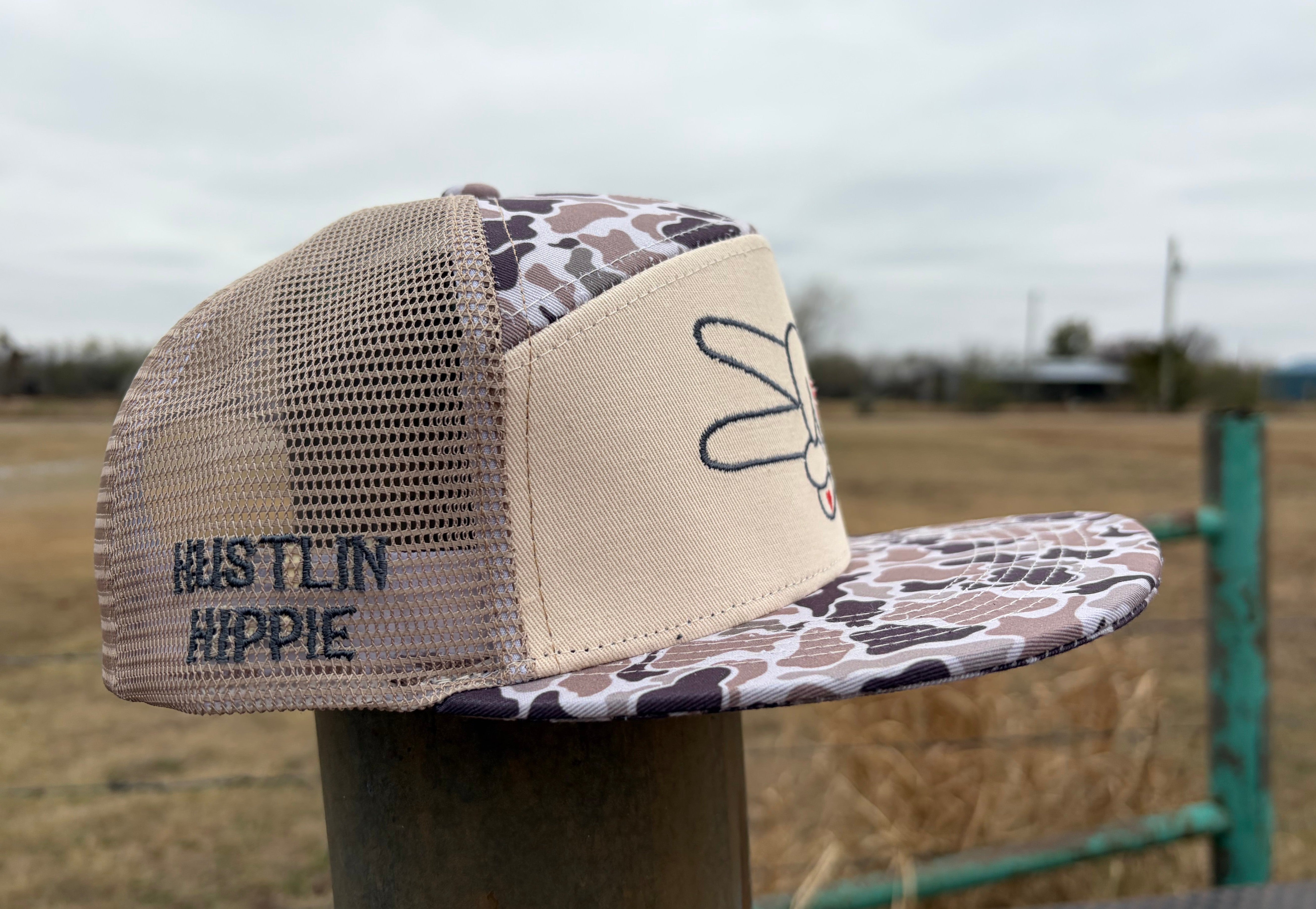 Side profile of Hustlin Hippie tan camo 7 panel mesh back trucker hat with embroidered side branding and outdoor-inspired camo pattern