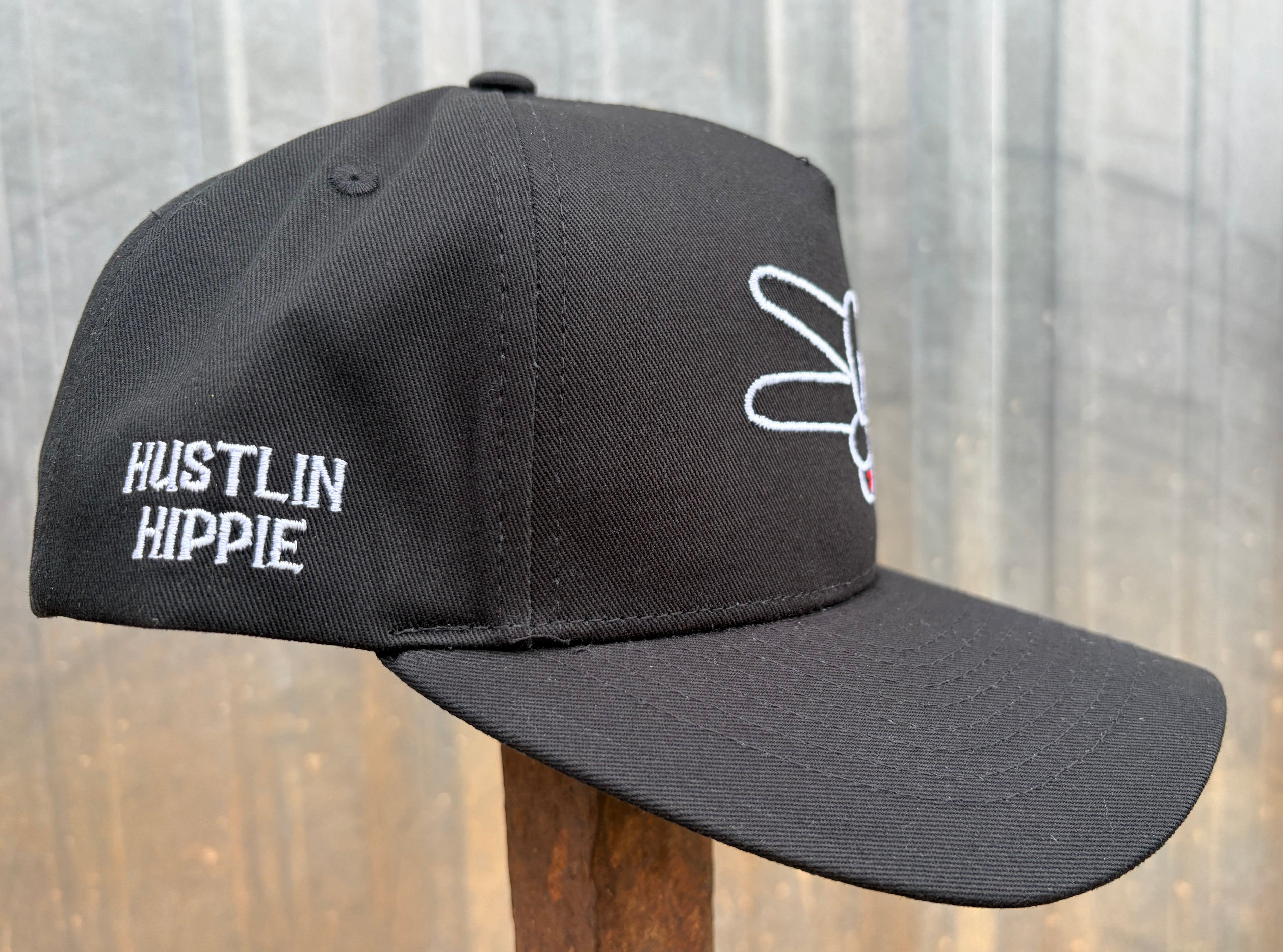 Side angle view of black Hustlin Hippie curved bill snapback hat featuring embroidered side logo and signature peace hand embroidery on structured front panel