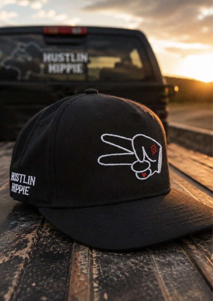 Black Hustlin Hippie signature curved snapback hat with embroidered peace hand logo resting in a pickup truck bed at sunset with golden hour lighting and Hustlin Hippie decal in background