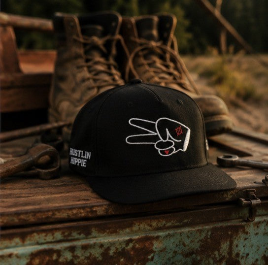 Black Hustlin Hippie curved snapback hat with embroidered peace hand logo displayed on a rock beside a lake at sunset with forest and water background outdoors
