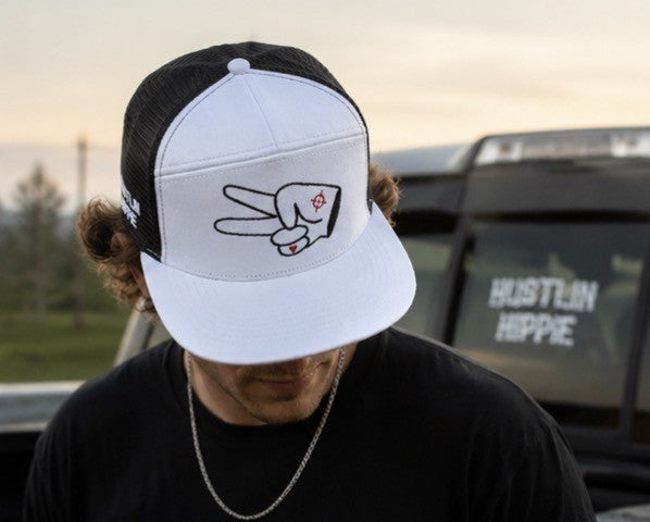 Person wearing a white and black Hustlin Hippie mesh trucker snapback hat with signature peace sign logo while standing in front of a pickup truck at sunset.