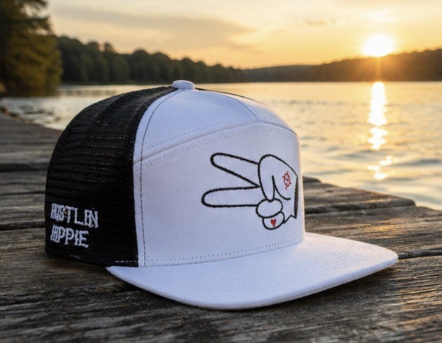 White and black Hustlin Hippie mesh trucker snapback hat with signature peace sign logo resting on a wooden dock beside a lake at sunset.