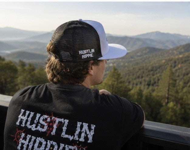 Person wearing a white and black Hustlin Hippie mesh trucker snapback hat overlooking a mountain landscape while wearing a Hustlin Hippie graphic tee.