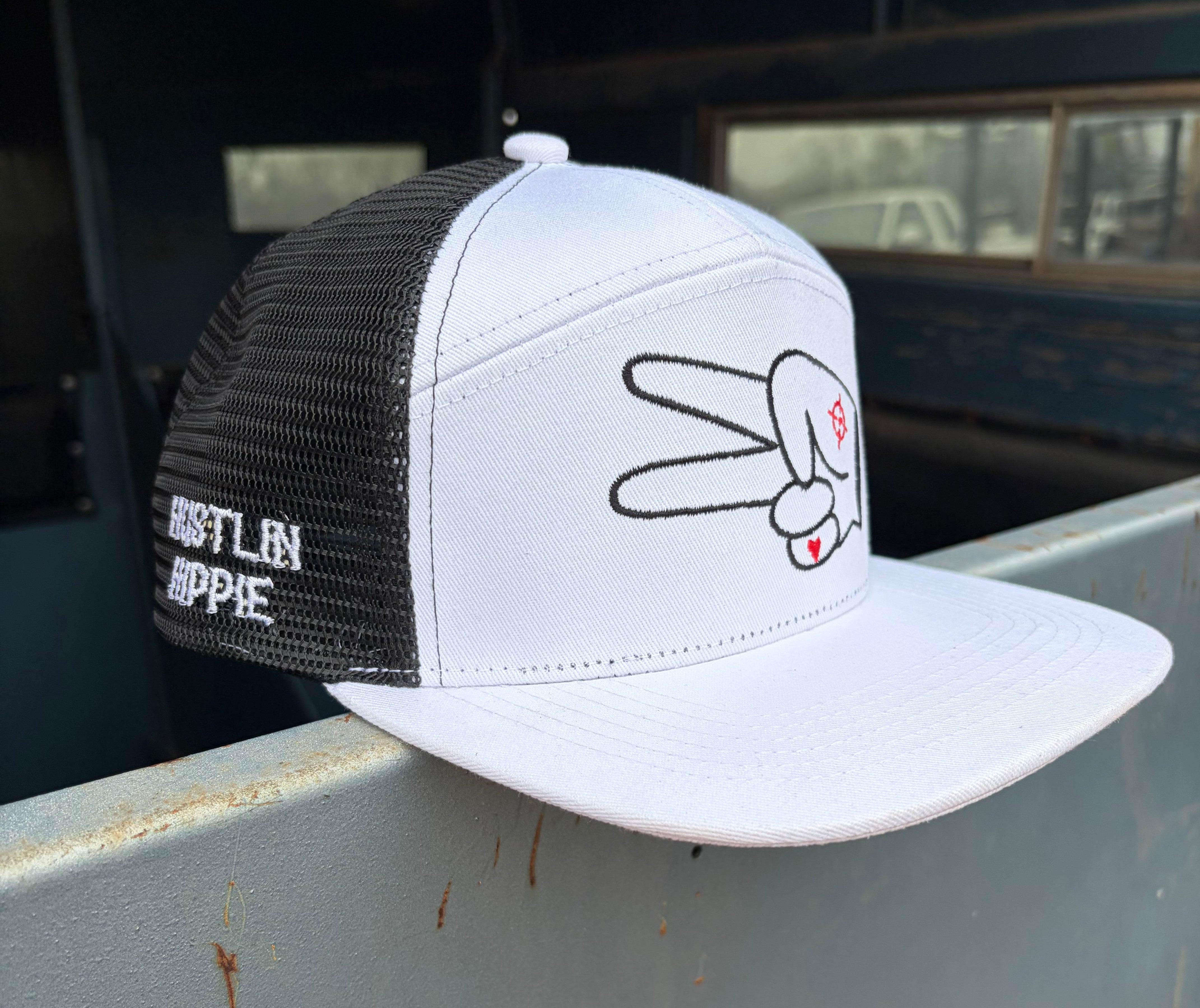 White and black Hustlin Hippie mesh trucker snapback hat with signature peace sign logo and embroidered side branding displayed on a rustic metal surface.