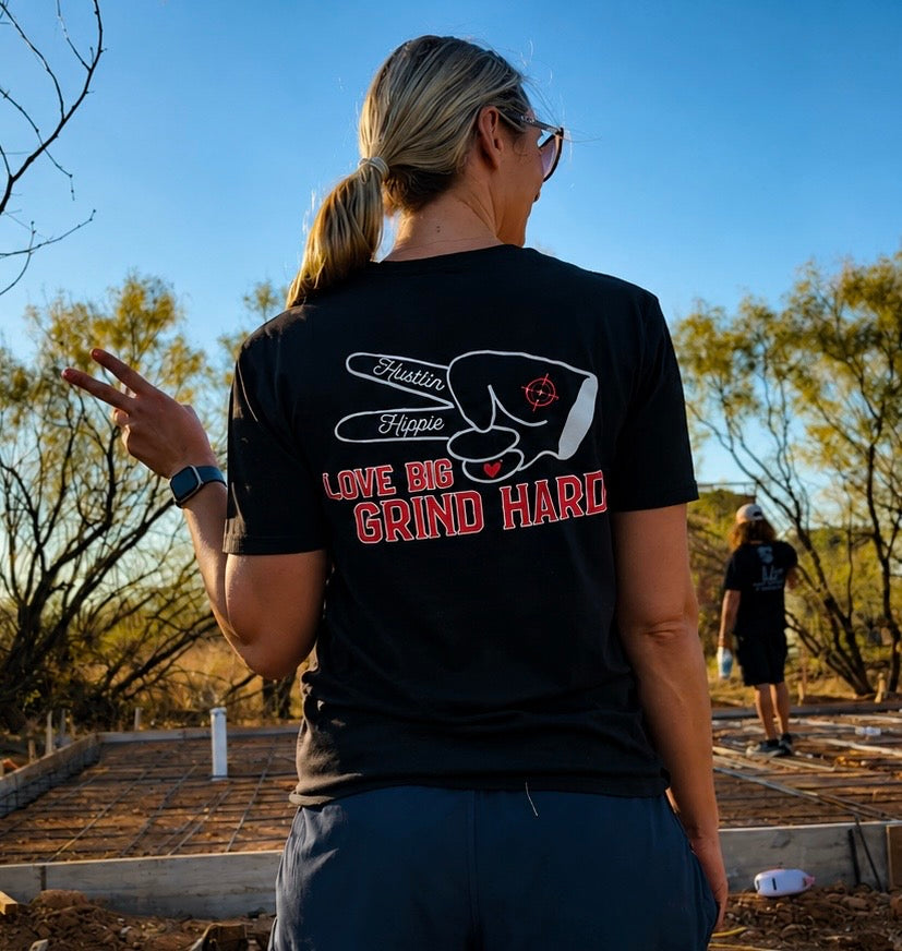 lifestyle photo of moan wearing black Hustlin Hippie Love Big Grind Hard graphic t-shirt at a construction site, featuring oversized back print with peace sign hand artwork and bold red lettering