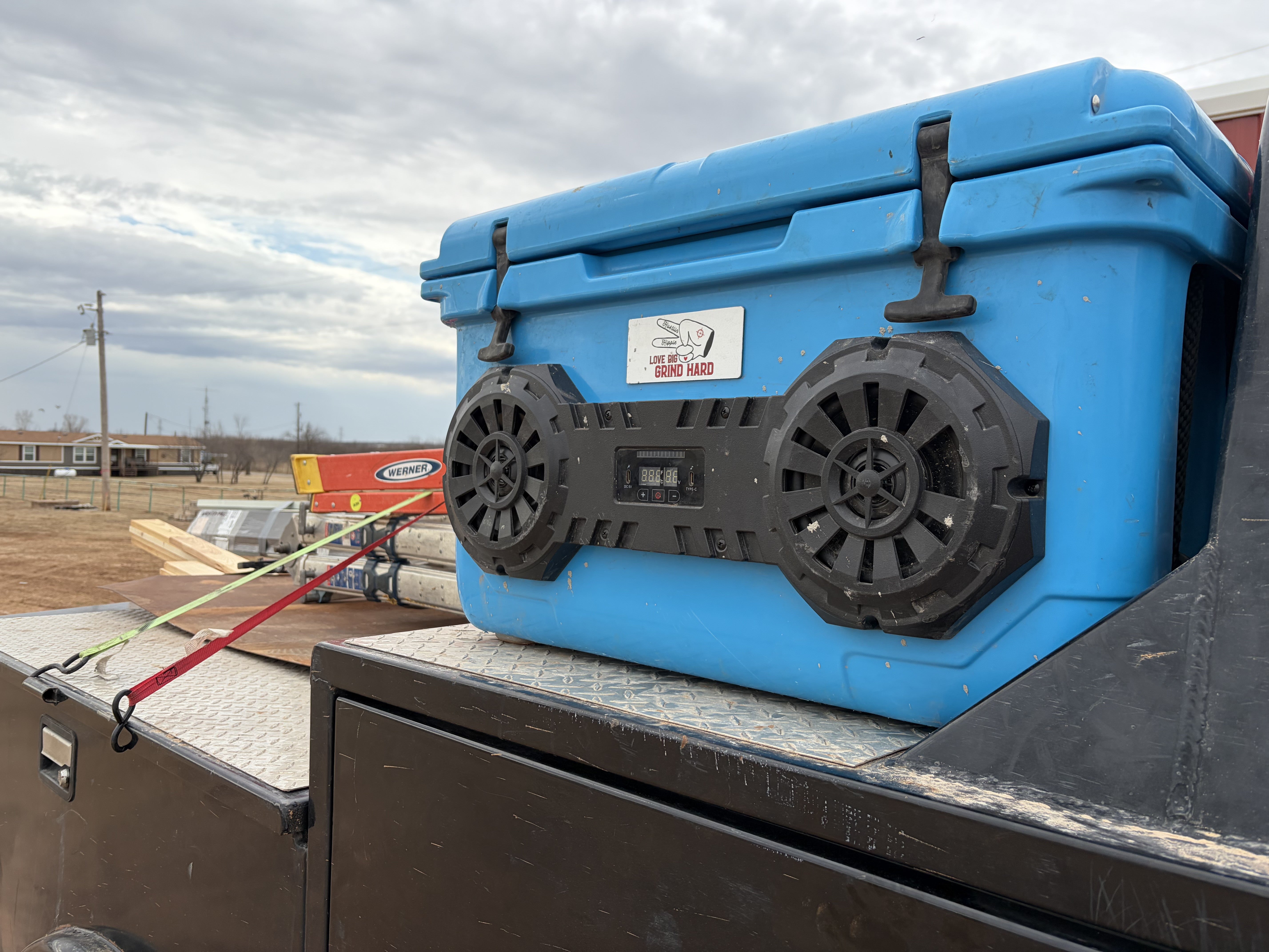 Blue Hound Boxx cooler with dual speakers on a truck bed against a cloudy sky.