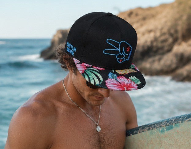 Man wearing a Hustlin Hippie Signature Floral Snapback Hat on the beach with surfboard during a tropical sunset surf session.