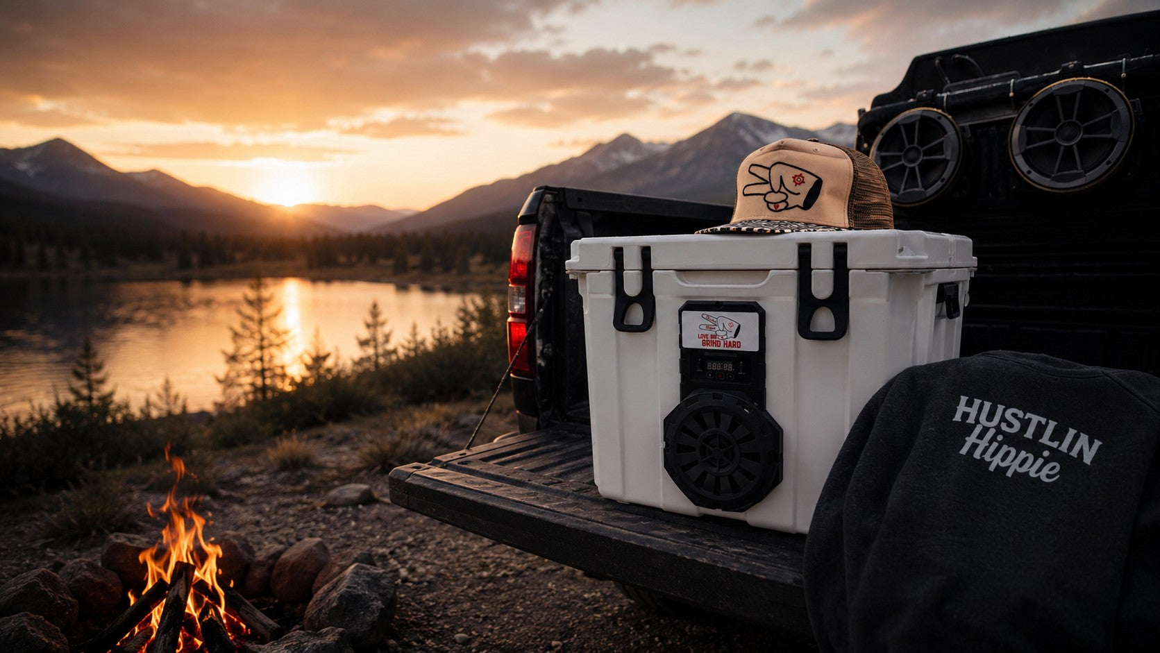 Hustlin Hippie outdoor lifestyle scene featuring a Hound Boxx Bluetooth cooler speaker, truck tailgate campfire setup, and adventure apparel at sunset