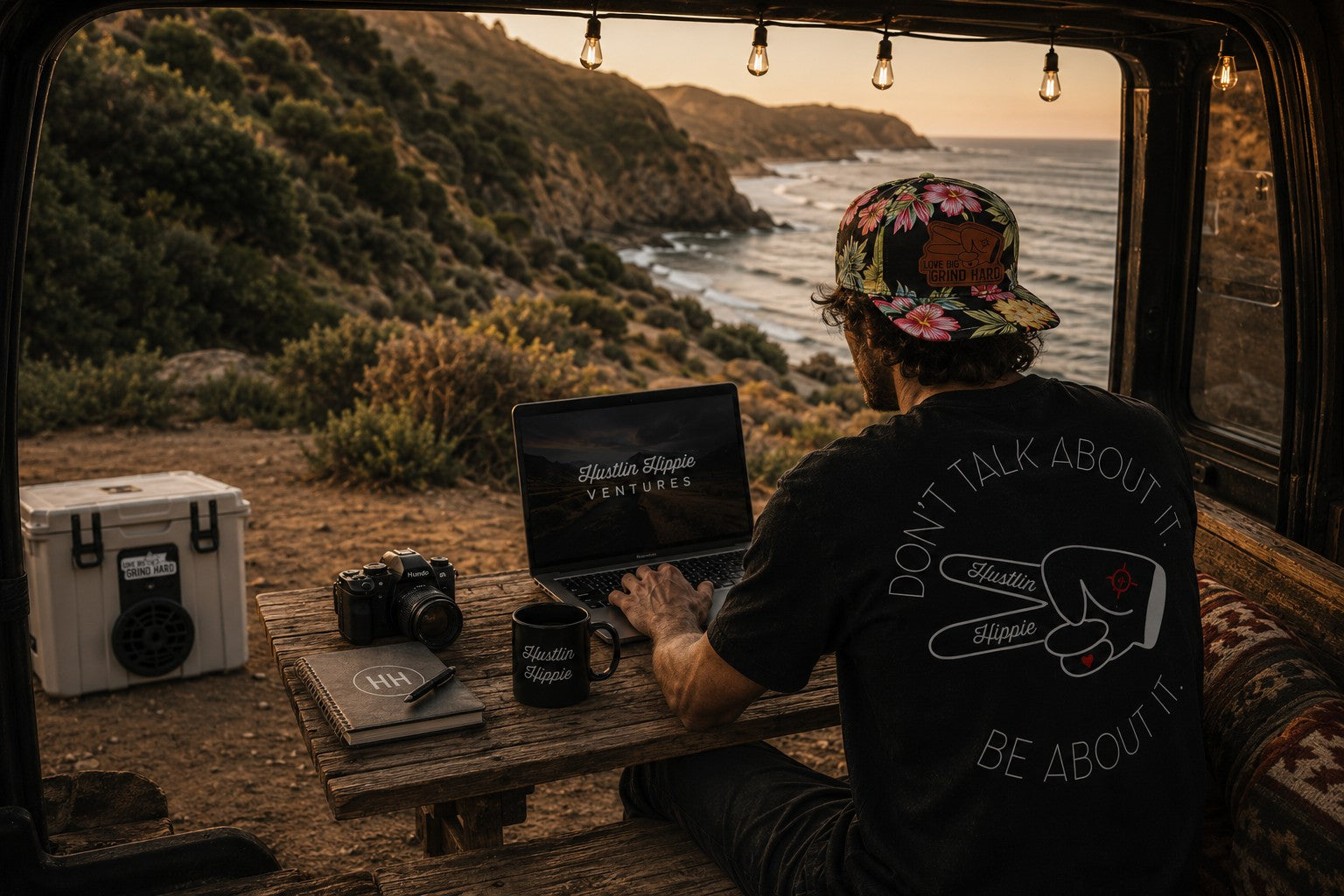 Man working from a camper van overlooking the ocean at sunset wearing Hustlin Hippie apparel and floral Love Big Grind Hard hat, blending work. travel, creativity and adventure lifestyle