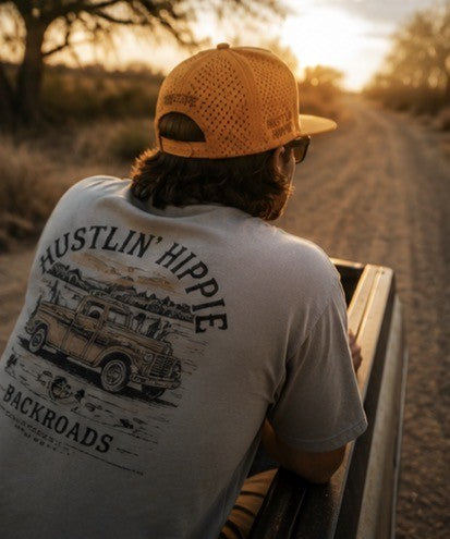 Person sitting in the back of a truck wearing a Hustlin Hippie hydro snapback while driving country backroads during golden hour.