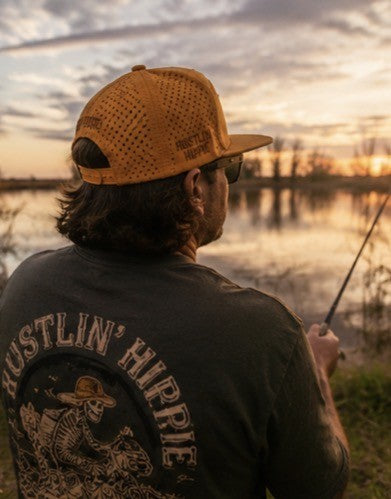 Person wearing a Hustlin Hippie hydro snapback while fishing at sunset beside the water with relaxed outdoor lifestyle vibes.