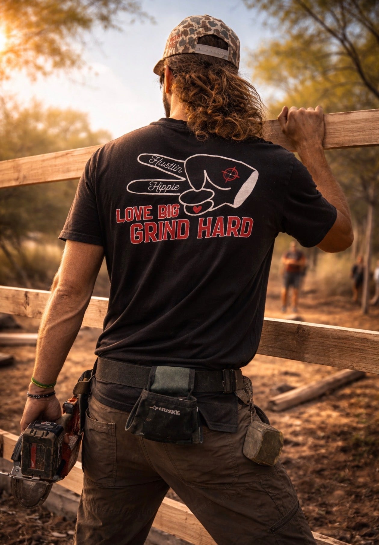 Man wearing black Love Big Grind Hard graphic t-shirt at outdoor construction site building wooden fence.