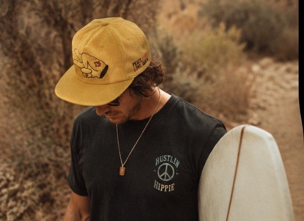 Man in a black Hustlin Hippie shirt wearing a mustard yellow Peace Love Hustle corduroy snapback hat holding a surfboard outdoors.