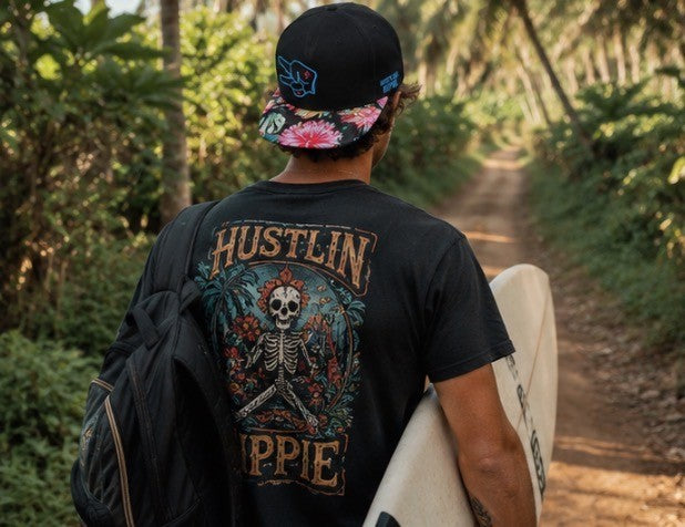 Back view of a surfer wearing a Hustlin Hippie tropical floral snapback hat while walking down a palm-lined trail with a surfboard.