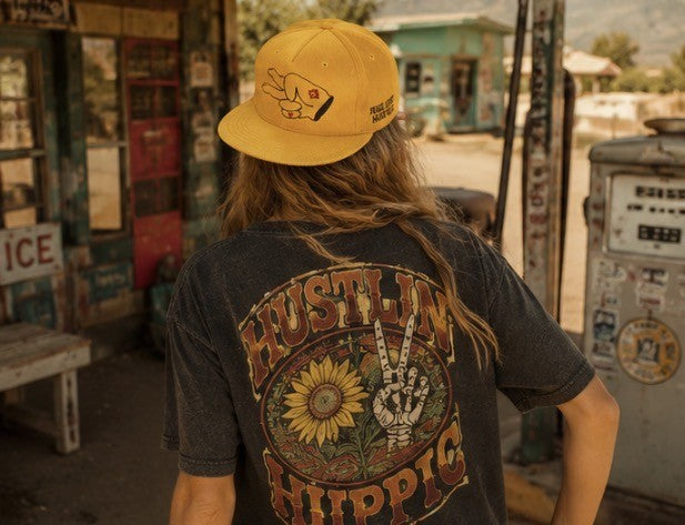 Woman wearing a mustard yellow Hustlin Hippie corduroy snapback hat at a vintage roadside gas station in a retro western setting.
