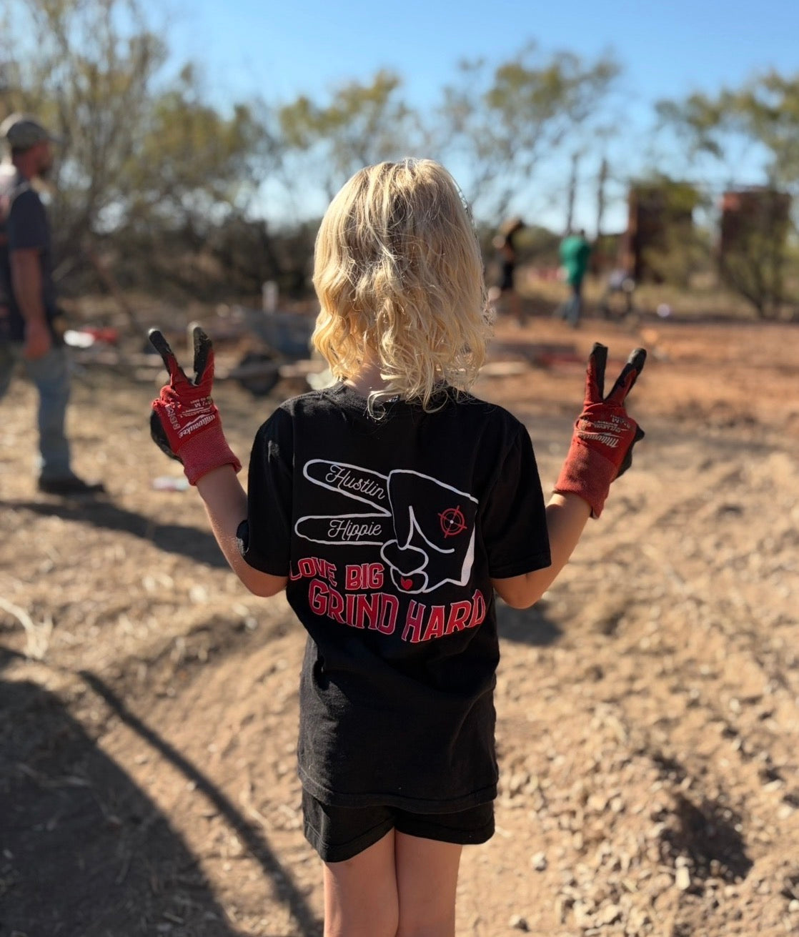 Child wearing black Hustlin Hippie Love Big Grind Hard youth t-shirt outdoors at a construction site.