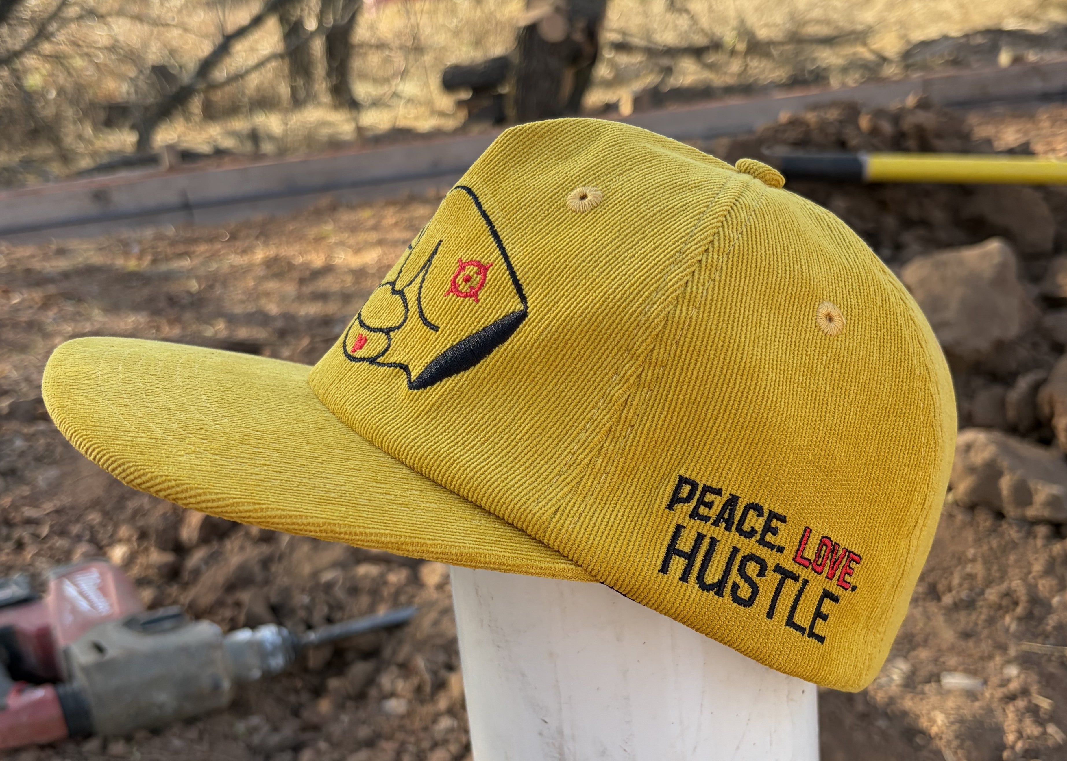 Mustard yellow corduroy snapback hat with PEACE LOVE HUSTLE embroidery and signature Hustlin Hippie logo outdoors.