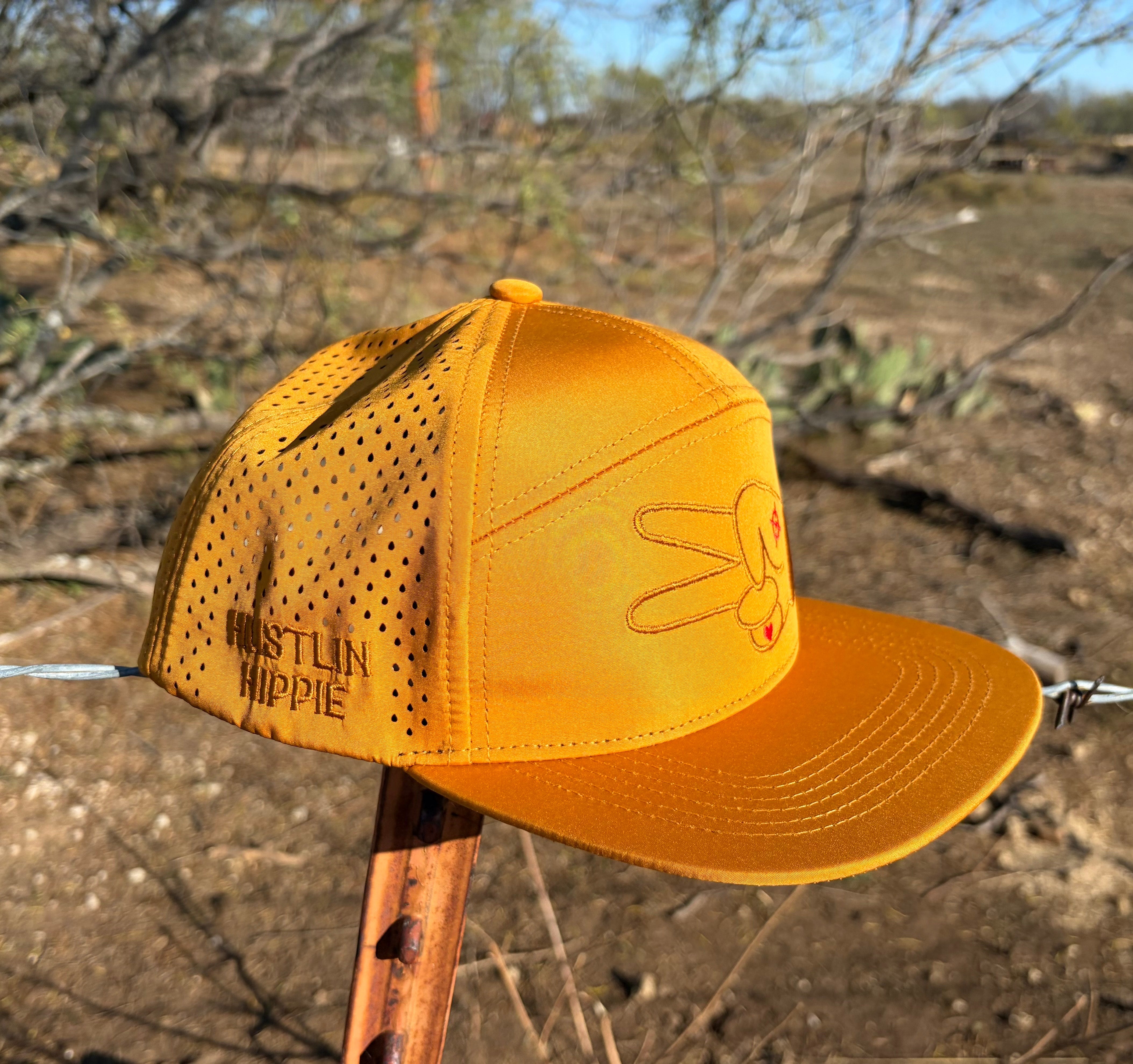 Side profile of Hustlin Hippie Signature Burnt Orange Hydro Snapback Hat featuring laser perforations and embroidered side logo outdoors in natural terrain