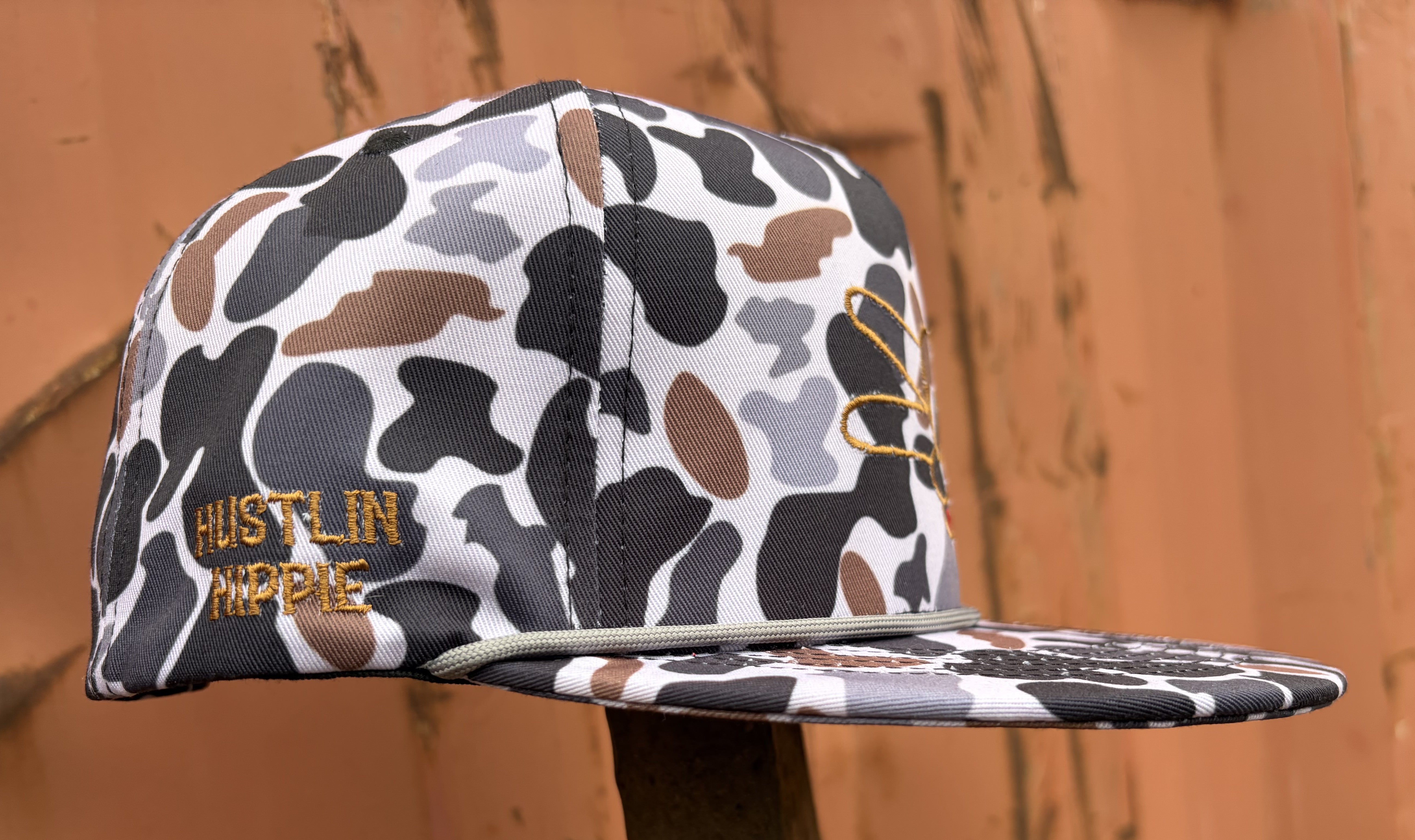 Hustlin Hippie Signature Midnight Camo rope snapback hat with embroidered logo and side branding shown at an angle against a rustic orange backdrop.