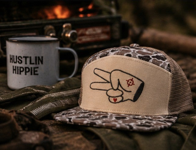 Hustlin Hippie Signature Tan Camo Trucker Hat beside campfire coffee mug at campsite outdoors