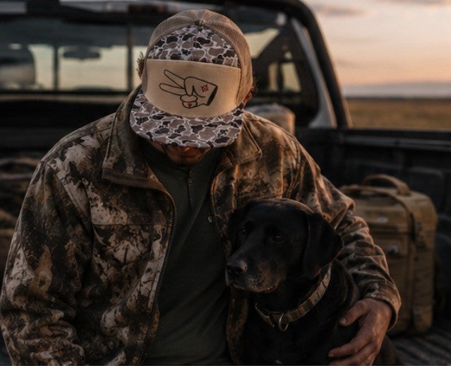 Hustlin Hippie Signature Tan Camo Trucker Hat styled with hunting gear and black dog in outdoor setting