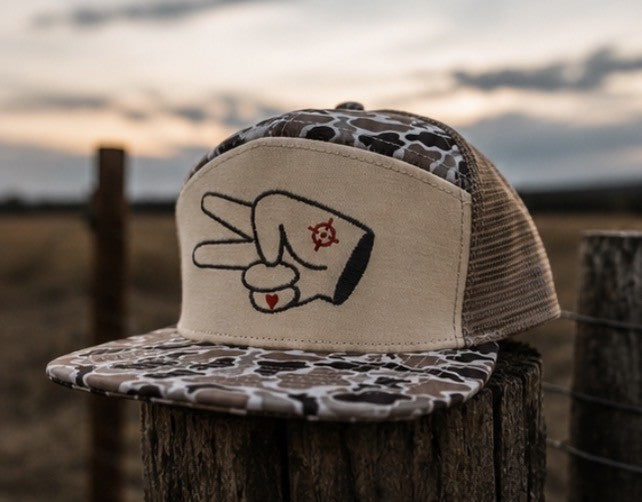Signature Tan Camo 7 Panel Mesh Back Trucker Hat resting on a rustic fence post in an outdoor country setting