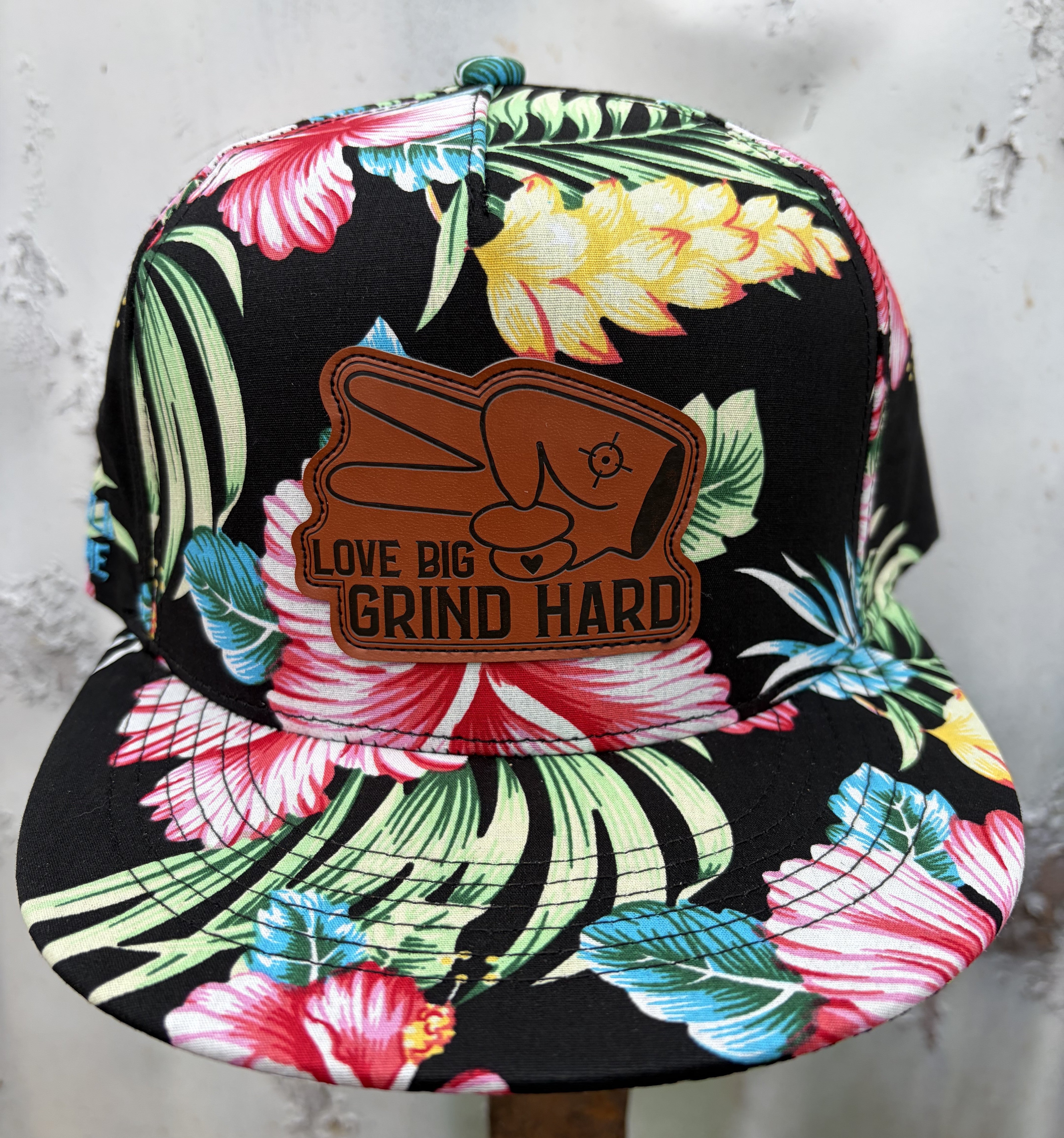 Front view of tropical floral snapback hat with leather Love Big Grind Hard patch and colorful hibiscus pattern.